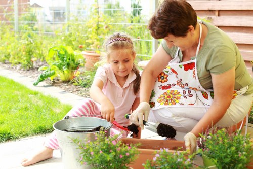 A woman and a young girl are engaged in gardening activities in a well-maintained backyard garden in Uxbridge. The woman, wearing a beige apron with a floral pattern, is kneeling on the paved patio area beside a rectangular flower bed filled with lush, green plants and flowering shrubs. The girl, dressed in a light pink top with pink and white striped leggings, is seated on the grass, actively involved in planting or potting, holding a small gardening tool. Surrounding them are vibrant green grass lawns, with a variety of garden elements visible including potted plants, garden soil, and a garden bucket. In the background, the garden showcases neatly arranged flower beds, and a wooden fence along the side of the property creates a private outdoor space. The scene is set under bright natural daylight, suggesting a clear, mild weather day, emphasizing the outdoor gardening environment that [Hedge Trimming Uxbridge] provides through professional garden maintenance services in the local area.