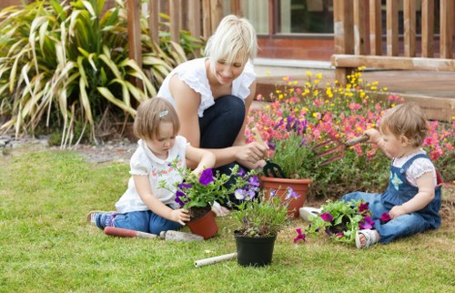 A cheerful woman with short blonde hair, dressed in a white sleeveless top and dark trousers, is kneeling on a well-maintained lawn in a garden in Uxbridge, engaging with two young children. The children, both girls with light brown hair, are sitting cross-legged on the grass, each tending to potted plants and flower arrangements. The garden features a lush green lawn, bordered by vibrant flower beds with pink, purple, and yellow flowers, and a backdrop of tall, leafy plants and a wooden deck with steps leading up to a house. Sunlight illuminates the scene under clear weather conditions, highlighting the natural textures of the grass, soil, and blooming flowers. The image captures a moment of outdoor gardening activity, emphasizing the importance of landscaping and garden maintenance services provided by Hedge Trimming Uxbridge, with a focus on creating attractive, family-friendly outdoor spaces in the local area.