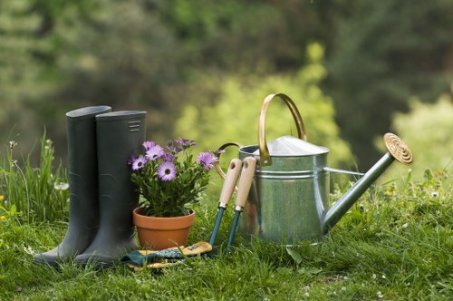 A neat, well-maintained garden scene featuring a line of tall, dark green rubber gardening boots placed upright on lush, green grass. To the right of the boots, there is a small terracotta flower pot filled with purple and white flowering plants, adding a touch of colour to the natural setting. Next to the pot, a pair of light wooden-handled garden shears rests on the grass, alongside other small gardening tools with colourful handles. Further right, a silver metal watering can with a curved handle and long spout is positioned, prepared for watering plants. The background shows a softly blurred, leafy garden environment with various green foliage, indicating a thriving outdoor space. The overall scene suggests outdoor gardening activities in a spacious, landscaped garden typical of gardens in Uxbridge and surrounding areas, with natural light suggesting a mild, pleasant day, perfect for garden maintenance by professionals such as Hedge Trimming Uxbridge.
